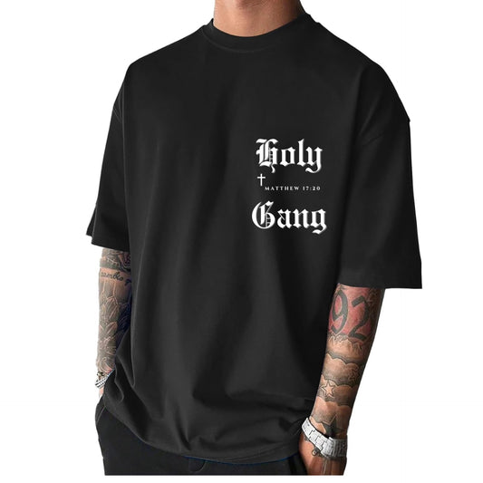 Front of male model wearing a black oversized Holy Gang t-shirt with white gothic logo, cross and Matthew 17:20 verse. 