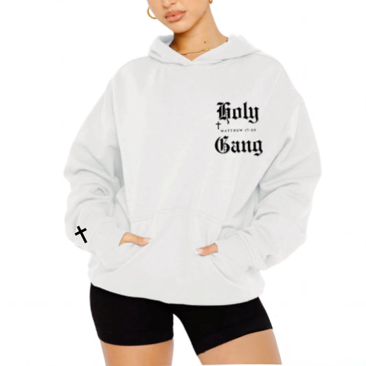 Woman wearing a white oversized hoodie with "Holy Gang"  printed in black gothic font on the left chest, paired with black shorts standing next to a plain background.