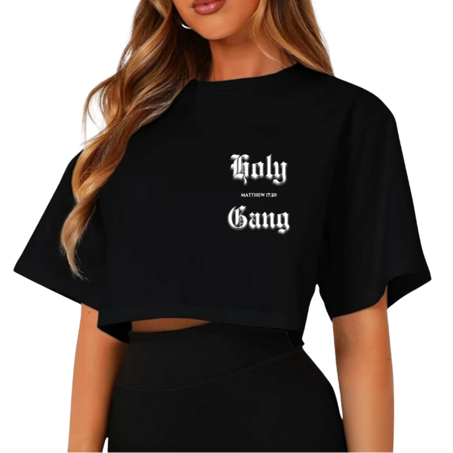 Front view of a woman wearing a black cropped t-shirt with the words "Holy Gang" printed in white Old English font, and the Bible verse reference "Matthew 17:20" between the words. The model has long wavy hair and is posing against a white background.
