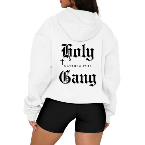 Back view of woman wearing a white oversized hoodie with "holy Gang" printed in large black gothic font, along with a small cross and bible verse reference "Matthew 17:20," styled with black shorts against a plain background. 