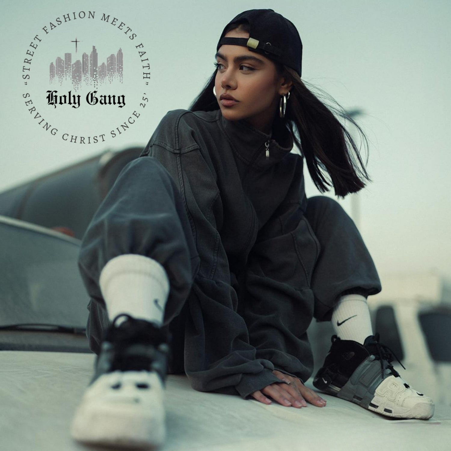 Young woman in dark gray streetwear outfit and black, white and gray Jordans crouching outdoors, wearing a backward cap, representing Holy Gang Christian streetwear brand with logo that says "Street Fashion Meets Faith - serving Christ since 25.'"